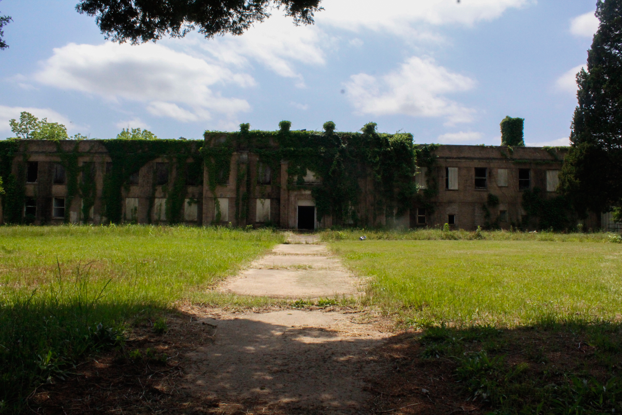 A concrete path leads across a grassy lawn to a large, abandoned two-story brick building covered — abandoned places photography in East Texas by Kaden Baker
