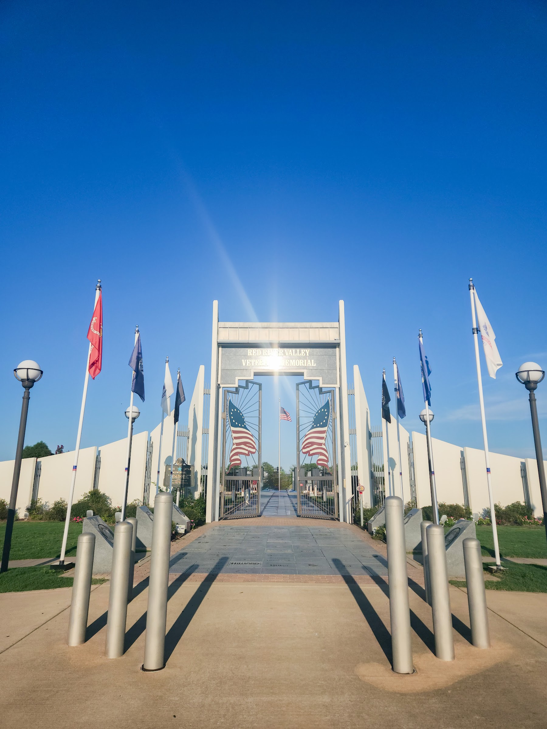 A metal archway entrance to a veterans memorial flanked by flagpoles and bollards casting long shadows under a — city photography in East Texas by Kaden Baker