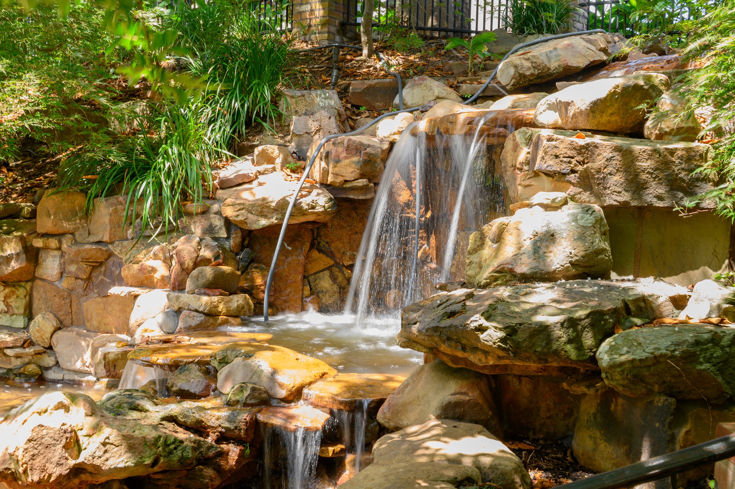 A small waterfall cascades down stacked golden-brown rocks into a pool, surrounded by lush green — city photography in Natchitoches, Louisiana by Kaden Baker