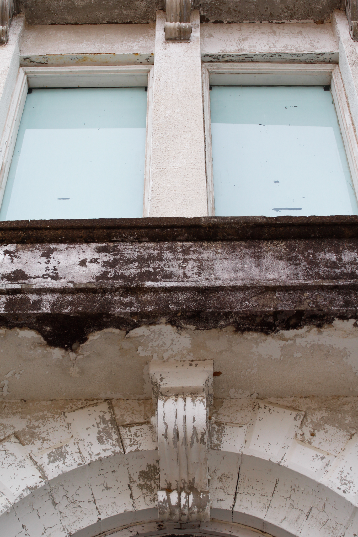 A low-angle view of a weathered white building facade featuring two pale blue windows above a peeling arch — city photography in East Texas by Kaden Baker