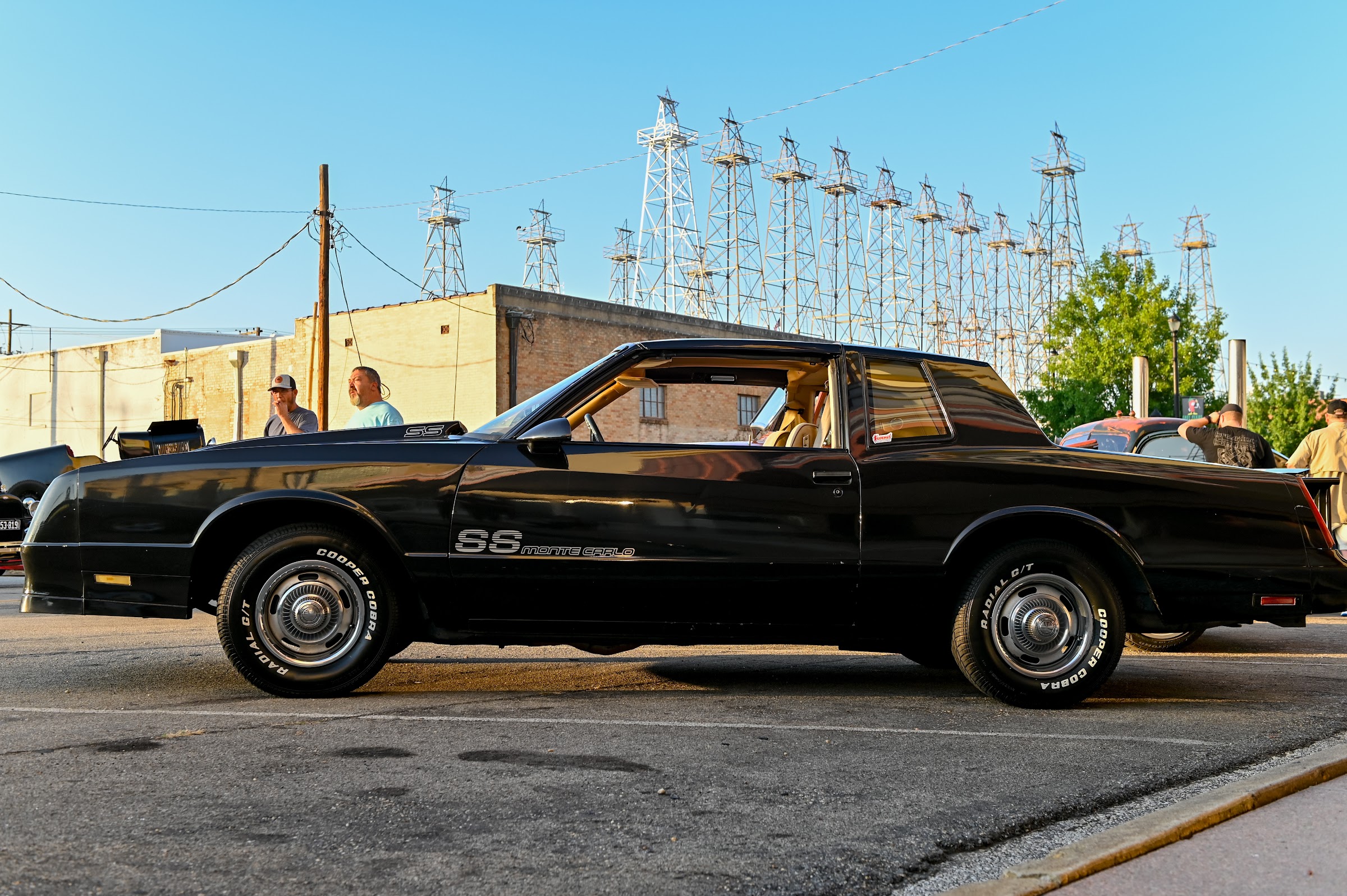 A black muscle car parked in front of historic steel oil derricks under a clear sky — car photography in East Texas by Kaden Baker