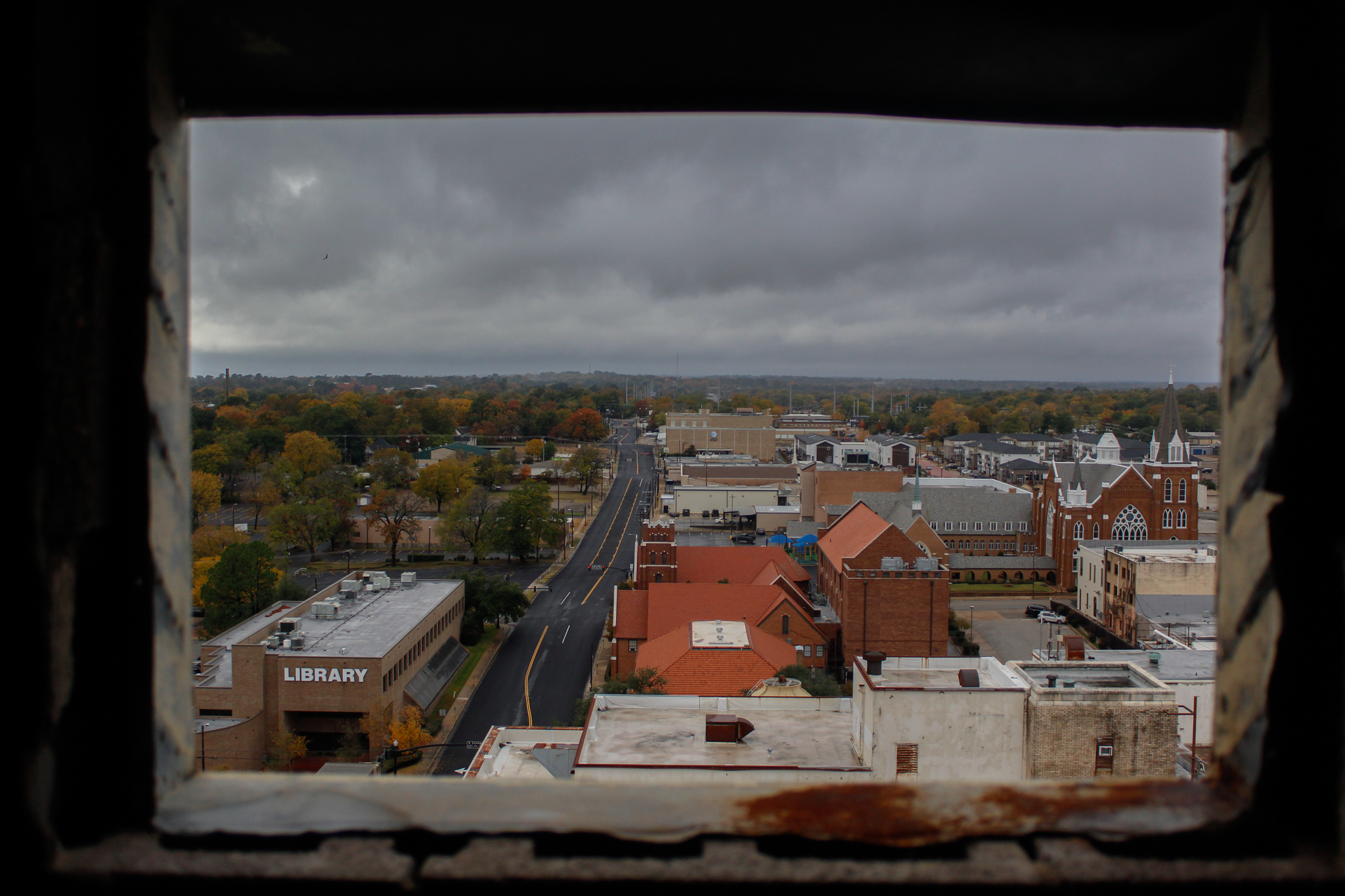 An elevated view framed by a dark window frame looking out over a small town street, brick church, and library — city photography in East Texas by Kaden Baker